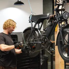 Man met blonde krullen voert mountainbike onderhoud uit. De fiets hangt aan een touw, klaar voor reparatie.