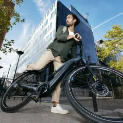 Man met rugzak haalt accu uit elektrische fiets. Moderne gebouwen en blauwe lucht op de achtergrond.