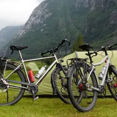 Twee fietsen staan klaar voor een fietsvakantie, met een tent en bergen op de achtergrond.