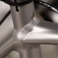 Close-up van de balhoofdbuis van een racefiets. Het frame is van aluminium, met een gladde, zilverkleurige afwerking. De kabels van de remmen lopen langs het frame.