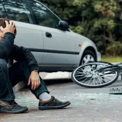 Man zit op stoeprand, hand in haar, na een fietser aangereden te zijn. Fiets ligt omgevallen, met glasscherven op straat.