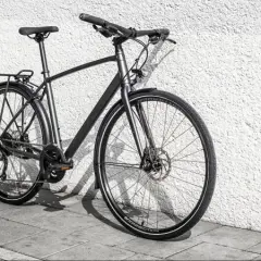 Grijze, moderne custom fiets met bagagedrager en schijfremmen, leunend tegen een witte, gestructureerde muur.