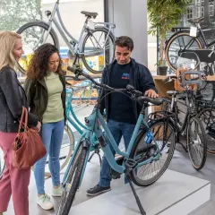 Een verkoper toont een lichtblauwe Gazelle bedrijfsfiets aan twee vrouwen in een fietsenwinkel.