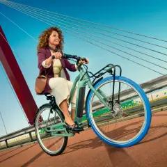 Vrolijke vrouw met krullend haar rijdt op een groene Swapfiets over een brug met blauwe lucht.