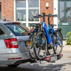 Twee fietsen, waaronder een blauwe elektrische fiets, hangen op een fietsendrager achter een zilveren auto. De fietsen steken uit, wat vragen oproept over hoe ver een fiets mag uitsteken op de auto.
