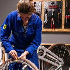 Vrouw in blauw werkpak haalt slag uit wiel. Kosten voor reparatie? Gereedschap hangt aan de muur.