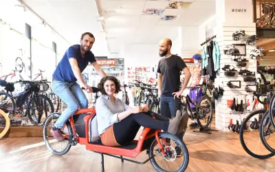 Een vrolijk stel test een rode bakfiets in een fietsenwinkel. De **oude fiets** is een blikvanger tussen de moderne racefietsen.