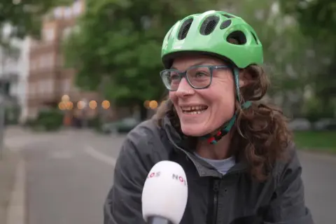 Vrolijke vrouw met groene fietshelm en bril, lachend in de camera. Ze vertelt over de fietshelm verplicht in Duitsland.