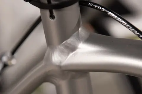 Close-up van de balhoofdbuis van een racefiets. Het frame is van aluminium, met een gladde, zilverkleurige afwerking. De kabels van de remmen lopen langs het frame.