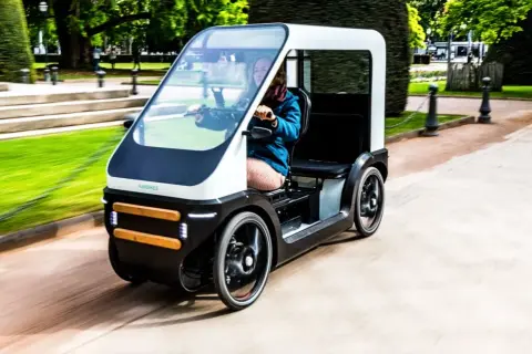 Een vrouw rijdt in een moderne fiets met dak door een park. De fiets heeft een strak design en biedt bescherming tegen het weer.