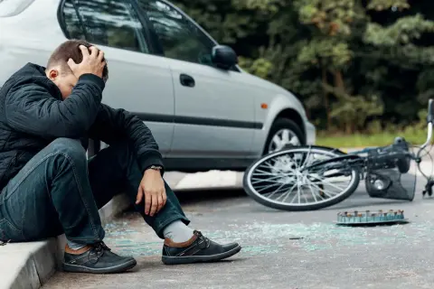 Man zit op stoeprand, hand in haar, na een fietser aangereden te zijn. Fiets ligt omgevallen, met glasscherven op straat.