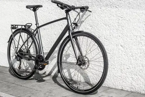 Grijze, moderne custom fiets met bagagedrager en schijfremmen, leunend tegen een witte, gestructureerde muur.