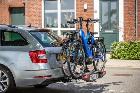 Twee fietsen, waaronder een blauwe elektrische fiets, hangen op een fietsendrager achter een zilveren auto. De fietsen steken uit, wat vragen oproept over hoe ver een fiets mag uitsteken op de auto.