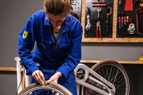 Vrouw in blauw werkpak haalt slag uit wiel. Kosten voor reparatie? Gereedschap hangt aan de muur.