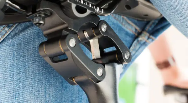 Gedetailleerde close-up van een fietszadel met een verende zadelpen afstellen. De mechanische delen zijn duidelijk zichtbaar, met een persoon in spijkerbroek op de achtergrond.