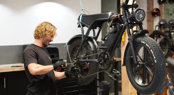 Man met blonde krullen voert mountainbike onderhoud uit. De fiets hangt aan een touw, klaar voor reparatie.