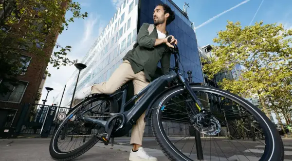 Man met rugzak haalt accu uit elektrische fiets. Moderne gebouwen en blauwe lucht op de achtergrond.