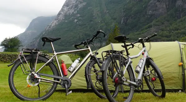 Twee fietsen staan klaar voor een fietsvakantie, met een tent en bergen op de achtergrond.