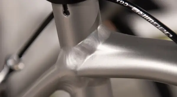 Close-up van de balhoofdbuis van een racefiets. Het frame is van aluminium, met een gladde, zilverkleurige afwerking. De kabels van de remmen lopen langs het frame.