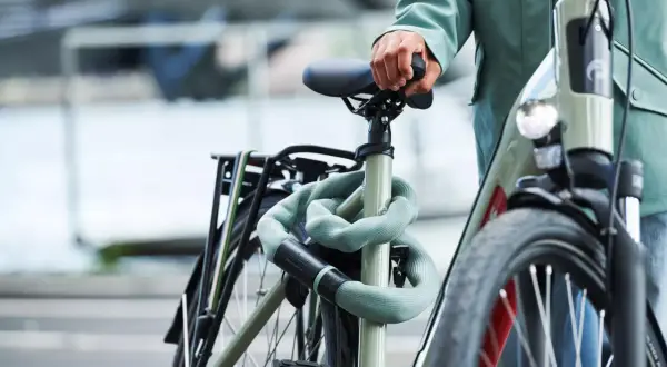 Een persoon met een groene jas houdt het zadel van een fiets vast. De fiets is beveiligd met een lichtgroene kettingslot. Denk aan een gps tracker fiets inbouwen voor extra veiligheid.
