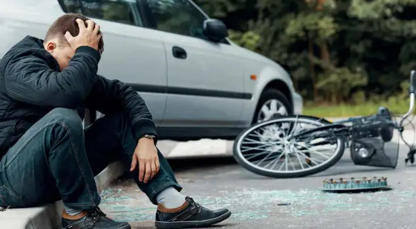 Man zit op stoeprand, hand in haar, na een fietser aangereden te zijn. Fiets ligt omgevallen, met glasscherven op straat.