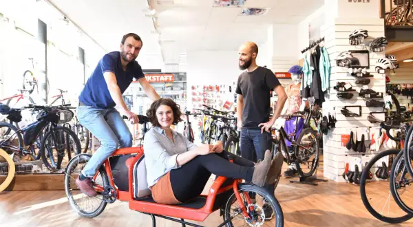 Een vrolijk stel test een rode bakfiets in een fietsenwinkel. De **oude fiets** is een blikvanger tussen de moderne racefietsen.