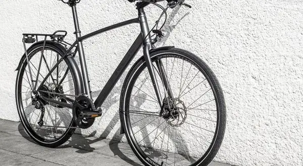 Grijze, moderne custom fiets met bagagedrager en schijfremmen, leunend tegen een witte, gestructureerde muur.