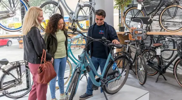 Een verkoper toont een lichtblauwe Gazelle bedrijfsfiets aan twee vrouwen in een fietsenwinkel.