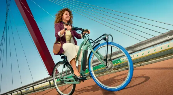 Vrolijke vrouw met krullend haar rijdt op een groene Swapfiets over een brug met blauwe lucht.