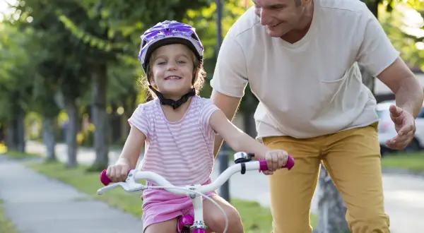 Een vader leert zijn dochter fietsen, met een helm op voor haar veiligheid. Zo zorgen we ervoor dat elk kind veilig op de fiets zit.