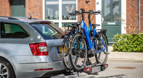 Twee fietsen, waaronder een blauwe elektrische fiets, hangen op een fietsendrager achter een zilveren auto. De fietsen steken uit, wat vragen oproept over hoe ver een fiets mag uitsteken op de auto.
