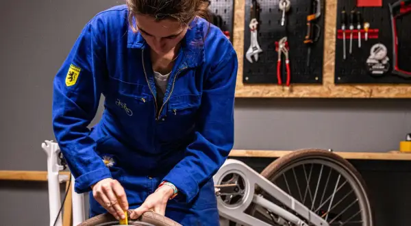 Vrouw in blauw werkpak haalt slag uit wiel. Kosten voor reparatie? Gereedschap hangt aan de muur.