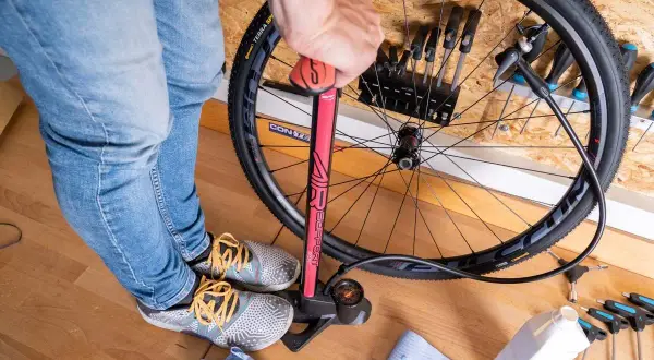 Een persoon pompt een tubeless band op met een rode fietspomp. Gereedschap en een flesje vloeistof liggen klaar.