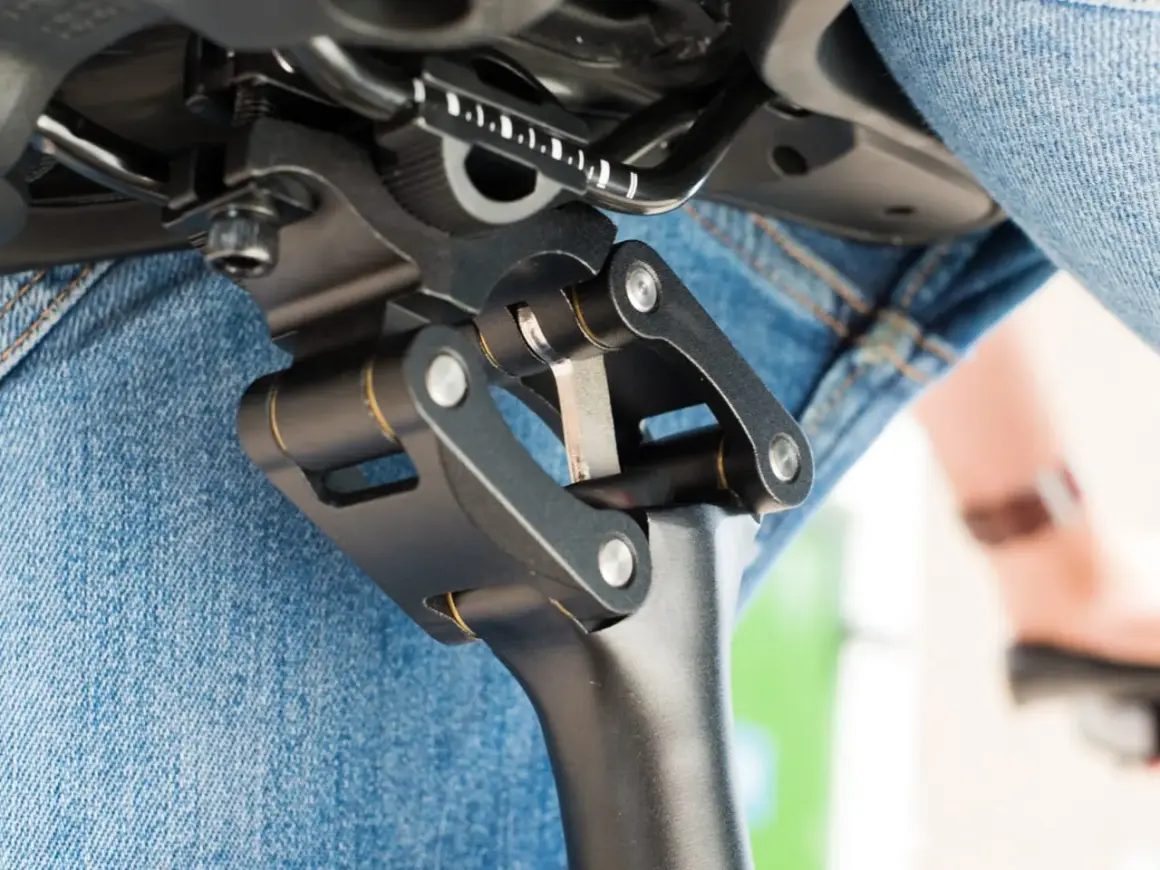 Gedetailleerde close-up van een fietszadel met een verende zadelpen afstellen. De mechanische delen zijn duidelijk zichtbaar, met een persoon in spijkerbroek op de achtergrond.