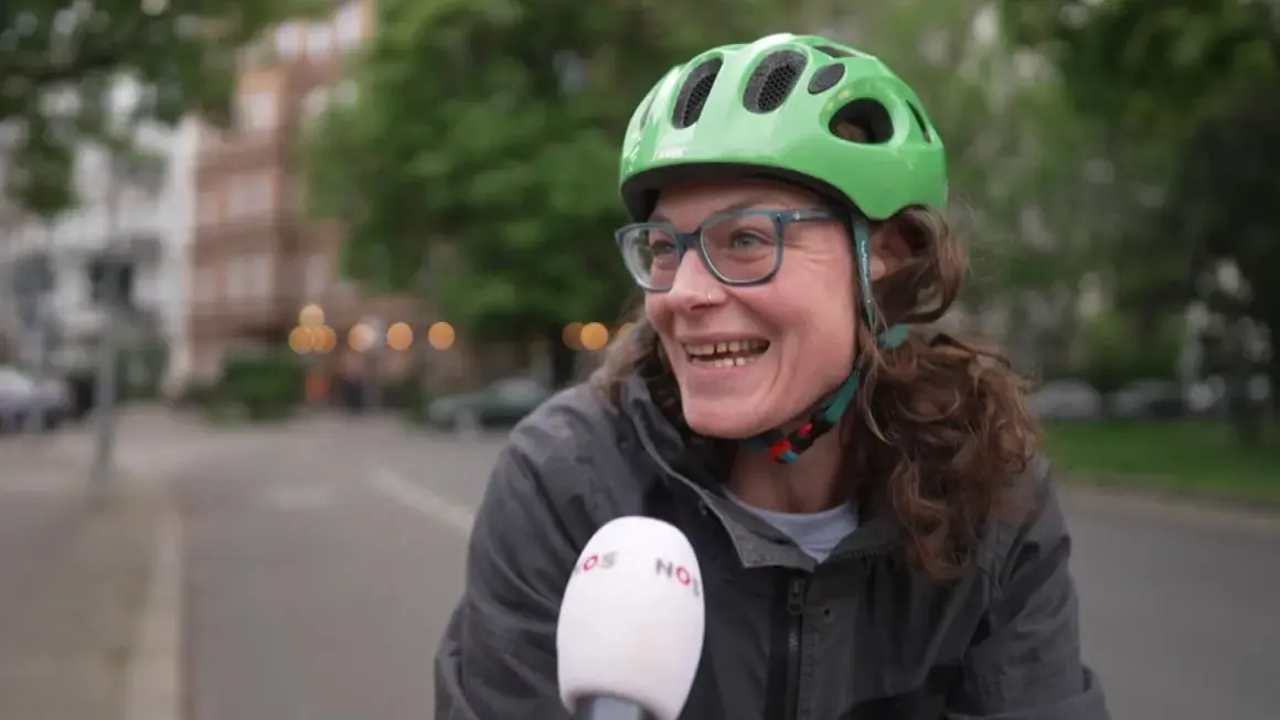 Vrolijke vrouw met groene fietshelm en bril, lachend in de camera. Ze vertelt over de fietshelm verplicht in Duitsland.