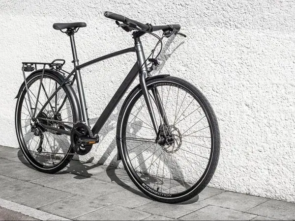 Grijze, moderne custom fiets met bagagedrager en schijfremmen, leunend tegen een witte, gestructureerde muur.