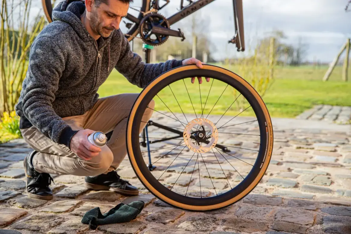 Man onderhoudt fietswiel met remschijf. Hij spuit met een busje, mogelijk voor het handrem fiets afstellen.