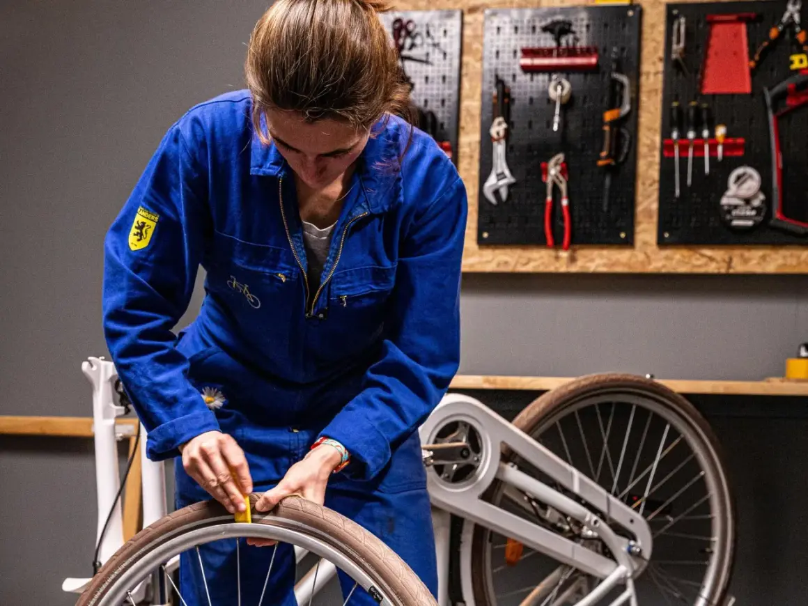 Vrouw in blauw werkpak haalt slag uit wiel. Kosten voor reparatie? Gereedschap hangt aan de muur.