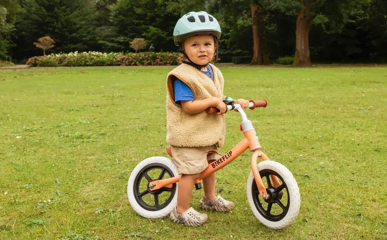 Een jong kind met helm en vestje op een loopfiets in het gras. Zo leren we elk kind veilig op de fiets.