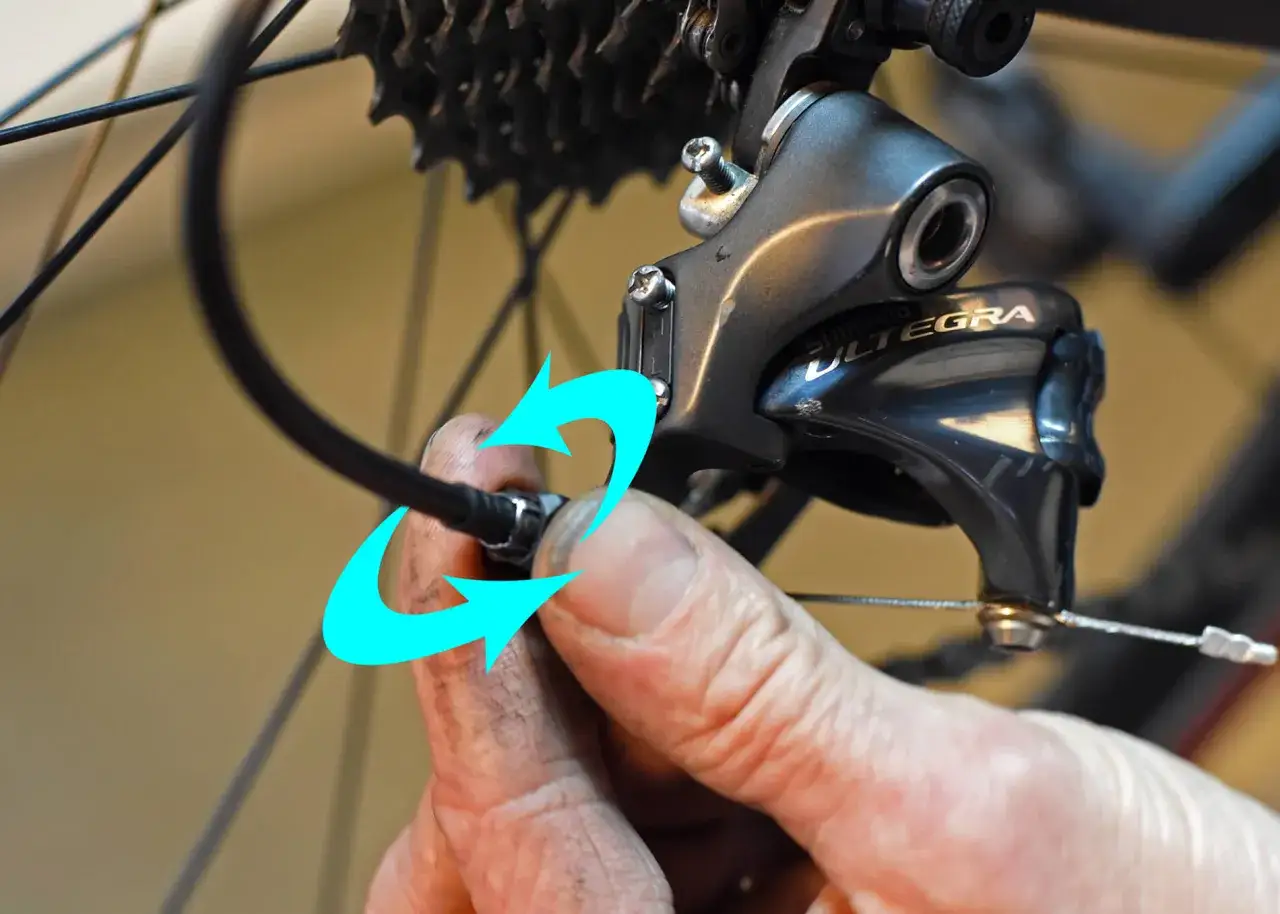 Hand draait aan kabel van derailleur. De ketting loopt van voorblad af, richting de cassette.