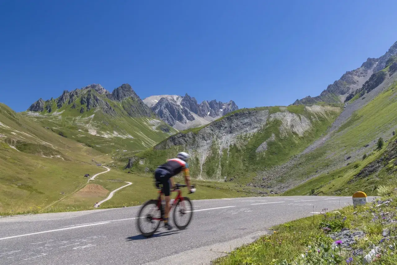 Fietser op een bergpas, omringd door groene hellingen en rotsachtige pieken. Een weg slingert door het landschap, waar misschien wel een marmotte te zien is.