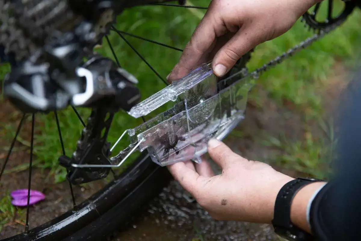 Handen met vuil gebruiken een kettingreiniger voor mountainbike onderhoud. De ketting wordt schoongemaakt met een transparant apparaatje.