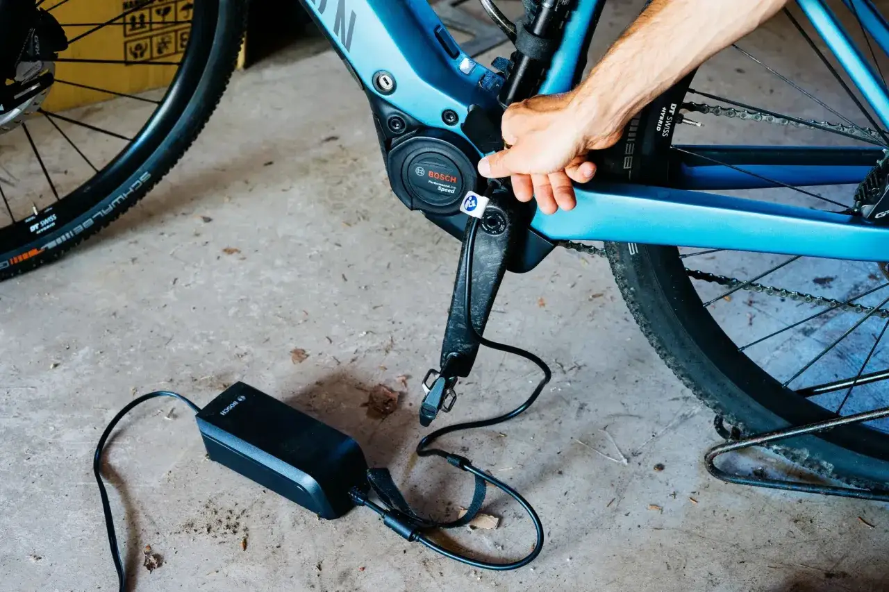 Hand sluit de oplader aan op de Bosch middenmotor van een blauwe e-bike. Klaar voor de volgende rit!