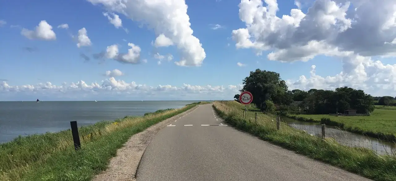 Een rustig asfaltpad langs het water, ideaal voor racefiets routes. Zeilboten op de achtergrond onder een blauwe lucht met witte wolken.