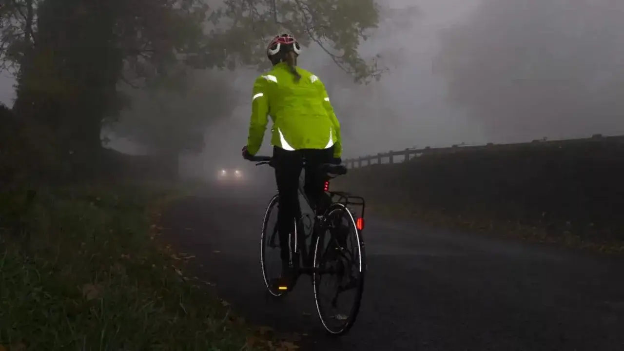 Een fietser met reflecterende kleding rijdt op een mistige weg. Koplampen van een naderende auto zijn zichtbaar. Zo ga je veilig op de fiets.