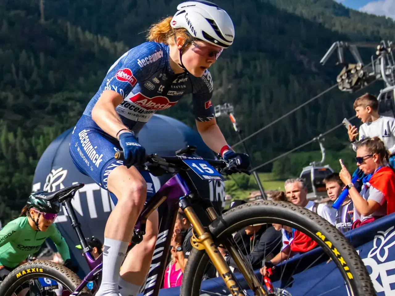 Nederlandse wielrenners strijden in een bergachtige omgeving. Een vrouw in een blauw tenue met 'Alpecin' en 'Deceuninck' logo's fietst met nummer 15.