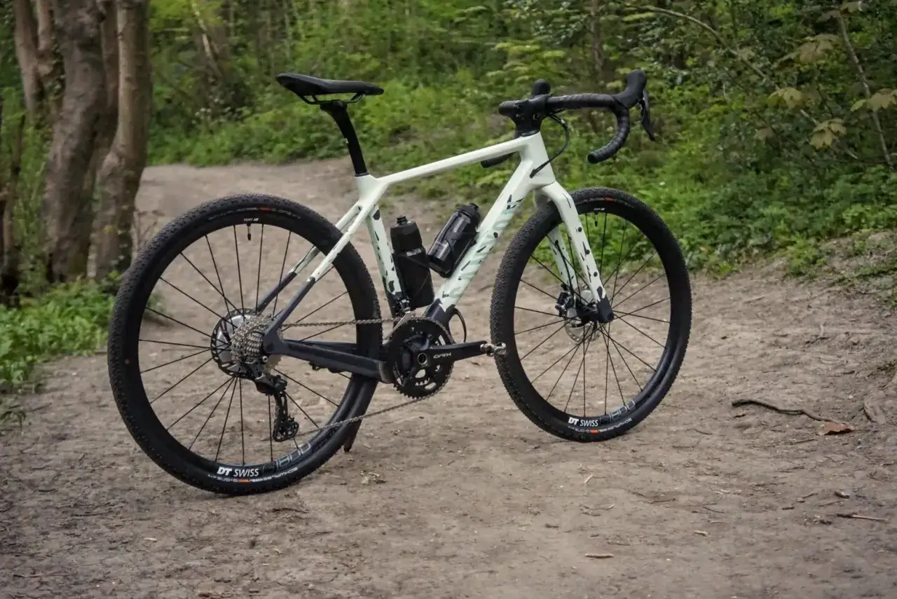 Lichtgroene cyclocross/gravel bike met brede banden en twee bidonhouders, geparkeerd op een zandpad in het bos.