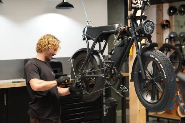 Man met blonde krullen voert mountainbike onderhoud uit. De fiets hangt aan een touw, klaar voor reparatie.