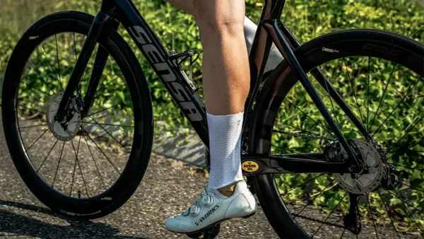 Een wielrenner met witte sokken en schoenen, op een racefiets met hoge velgen. De fiets is zwart met een Sensa frame.