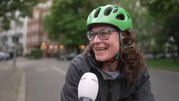 Vrolijke vrouw met groene fietshelm en bril, lachend in de camera. Ze vertelt over de fietshelm verplicht in Duitsland.